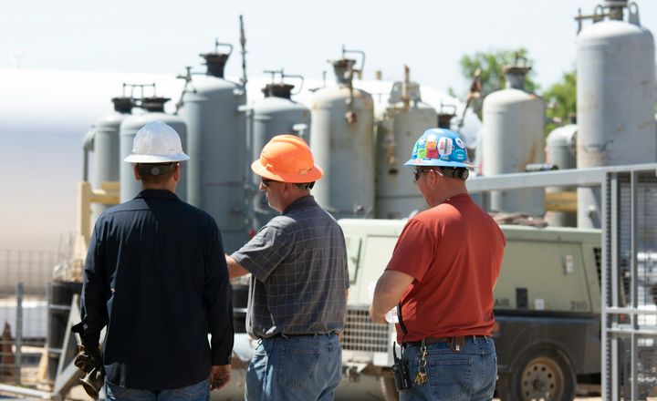 Three men in hard hats walking past the tanks of an oil field facility